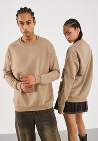 To modeller med flettet hår iført beige hettegensere; en vender seg mot kameraet med hendene foldet, den andre snur seg litt bort, iført skjørt og støvler.