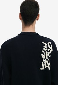 Marineblå strikket sweater med ribbet halsudskæring og hvid grafisk tekst på ryggen. Løs pasform med et struktureret mønster.