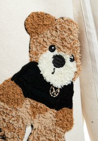 Tela beige con un diseño de oso de peluche en texturas marrón y blanca. El oso lleva una camiseta negra con un emblema circular como acento.