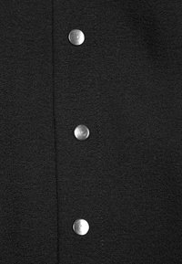 Tissu noir texturé avec trois boutons-pression en argent brillant alignés verticalement sur une couture centrale.