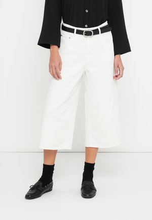 Pantaloni bianchi a gamba larga, tagliati sopra la caviglia, con una texture liscia, abbinati a una cintura nera e mocassini neri, indossati con calzini neri alti.