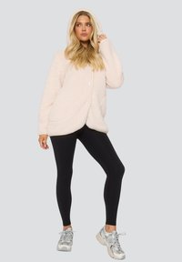 Lichtroze oversized capuchonjack gemaakt van fluffy stof, met knopen en een ronde zoom, gecombineerd met zwarte legging en grijze sneakers.