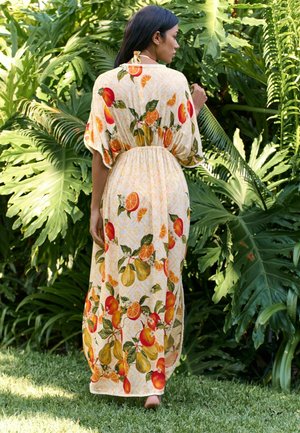 Žena s dlhými tmavými vlasmi kráča naboso po tráve, oblečená v dlhých krémových šatách s farebnou potlačou citrusového ovocia, obklopená veľkými zelenými listami.