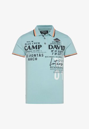 Polo shirt azzurro chiaro con testo stampato in bianco e nero, strisce arancioni a contrasto sul colletto e una texture morbida in cotone.