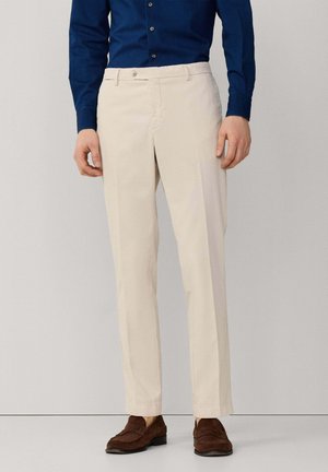 Mann trägt beige Hose, dunkelbraune Loafers und ein dunkelblaues, langärmeliges Hemd mit Knöpfen, steht vor einem hellen Hintergrund.