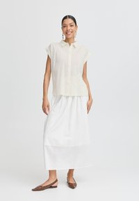 Camicia bottonata gialla chiaro a maniche corte con righe verticali, abbinata a una gonna bianca fluida lunga fino alle caviglie e scarpe flat slingback marroni.