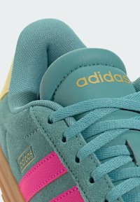 Turkos mocka sneakers med rosa ränder, guld "adidas"-logotyp, texturerad meshfoder och hållbar gummisula. Snörning för en säker passform.