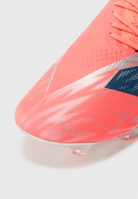 Scarpa da calcio rosa brillante con superficie testurizzata, dettagli in argento e un elemento di design blu scuro. Presenta una forma elegante e tacchetti trasparenti.