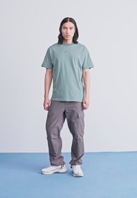 Jovem com cabelo comprido vestido com uma t-shirt verde da Nike, calças cargo cinzas e ténis brancos, em pé sobre um chão azul contra uma parede branca.