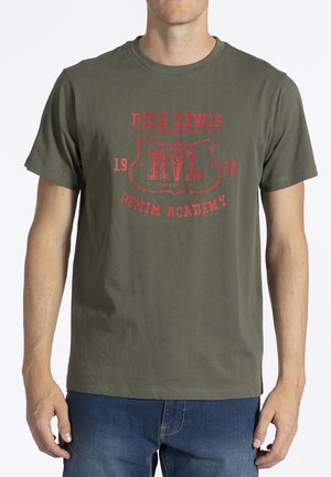 Homme portant un T-shirt vert olive avec le logo rouge "Rica Lewis Denim Academy" et un jean bleu, debout devant un fond blanc.
