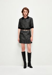 Grijze tweed cropped jacket met zwarte kraag en manchetten, gecombineerd met een bijpassende mini-rok met franje, zakken en zilveren accenten. Zwarte kniehoge laarzen.