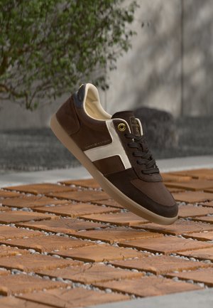 Καφέ και λευκό αθλητικό παπούτσι Tommy Hilfiger με μαύρα κορδόνια που αιωρείται πάνω από ξύλινο πλακάκι δάπεδο με φυτά και γκρι τοίχο στο φόντο.