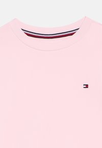 Svetloružové tričko s okrúhlym výstrihom, s malým obdĺžnikovým logom v červenej, bielej a námorníckej modrej farbe na hrudi a prúžkovaným detailom goliera na vnútornej strane výstrihu.