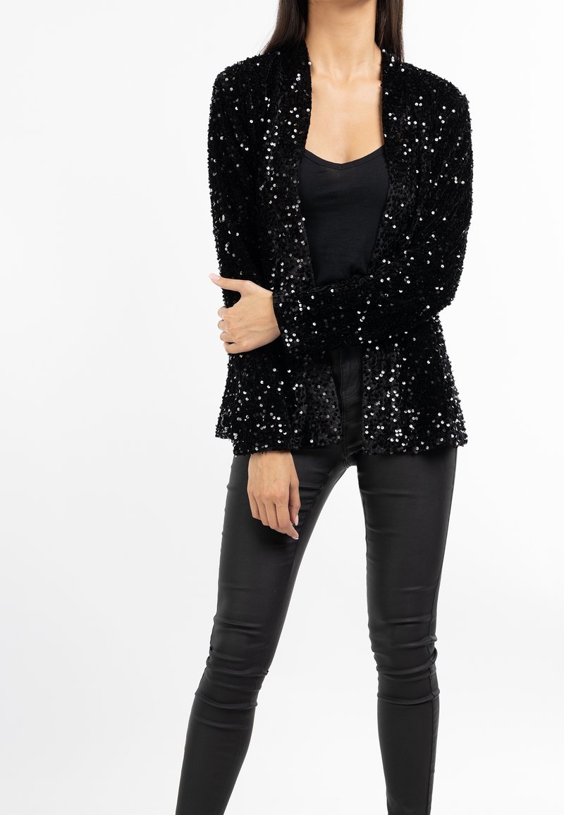 Blazer nero in paillettes con superficie testurizzata, maniche lunghe, design aperto sul davanti, abbinato a pantaloni in pelle nera.