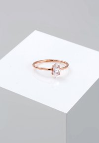 Anello in oro rosa con una pietra trasparente a taglio marquise, esposto su un piccolo piedistallo geometrico bianco in un ambiente minimalista.