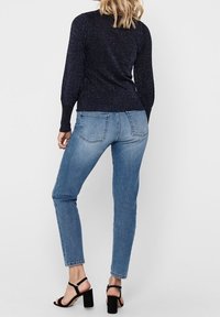 Blonde Frau trägt einen dunkelblauen Strickpullover, blaue Slim-Fit-Jeans und schwarze Blockabsatzsandalen und steht mit dem Rücken zur Betrachterin.