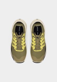 Ζευγάρι από αθλητικά παπούτσια σε ελαιδί πράσινο και κίτρινο με μπεζ σόλες, φωτισμένα από πάνω, πάνω σε λευκό φόντο.