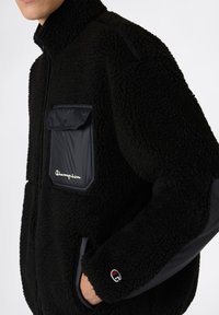 Chaqueta de forro polar negra con cuello con cremallera, material texturizado, un bolsillo en el pecho con cremallera y detalles de logotipo en el bolsillo y la manga.