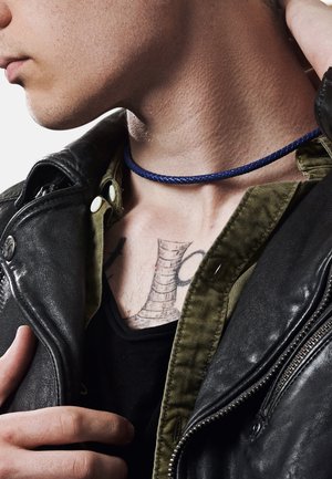 Schwarze Lederjacke über einem olivgrünen Hemd; blauer geflochtener Kordel-Choker; sichtbares Tattoo auf der Brust; Nahaufnahme von Hals und Schulter.
