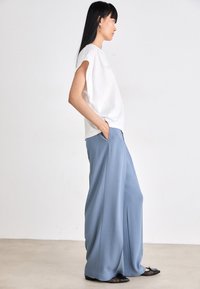 Haut sans manches blanc associé à un pantalon léger bleu clair, ample et doté de poches latérales avec une texture lisse. Des chaussures plates noires viennent compléter le look.