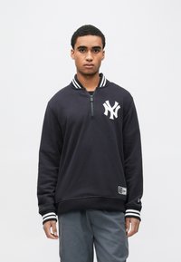 Tengerészkék pulóver félcipzárral, csíkos gallérral és bordázott mandzsettákkal. A mellrészen fehér logóval, az alján pedig egy kis címkével díszített.