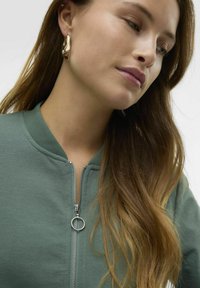 Sweatshirt vert à fermeture éclair avec col côtelé et détail de fermeture éclair argenté. Porté avec des boucles d'oreilles en or et présente des cheveux longs et ondulés.