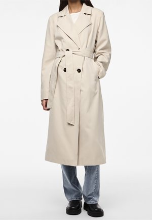 Mujer con trench coat beige con cinturón, jeans azules y zapatos negros, de pie con una mano en el bolsillo, sobre un fondo blanco liso.
