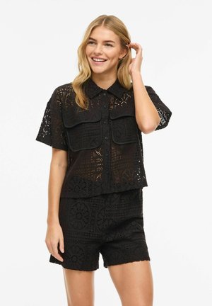 Uśmiechnięta kobieta z długimi blond włosami, ubrana w czarną koronkową koszulę z krótkimi rękawami, kieszeniami z klapkami i pasujące szorty z wysokim stanem.