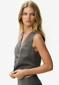 Szary top bez rękawów w prążki, z dekoltem w kształcie litery V, czterema guzikami z przodu i dopasowanym krojem. Tkanina ma gładką fakturę.