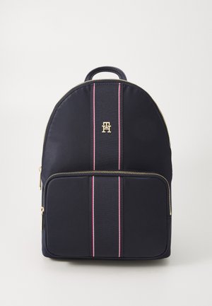 Sac à dos bleu marine avec poche zippée à l'avant, fermeture éclair dorée, détail en tissu rayé verticalement et petit logo doré en haut au centre.