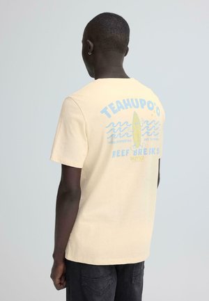 Homme portant un t-shirt jaune pâle avec un graphisme de planche de surf bleue et jaune et le texte « TEAHUPO'O REEF BREAKS » dans le dos, tourné de dos.