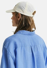 Lichtblauw linnen shirt met een achterste beleg, gedragen onder een crèmewitte baseballcap. De cap heeft een klein logo aan de voorkant. Haar is los en golvend.