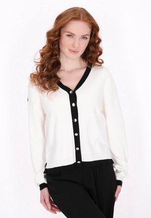 White cardigan with black trim, featuring a V-neck and gold buttons down the front. Long sleeves with ribbed cuffs and a textured knit.