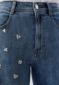 Jean en denim bleu avec des ornements métalliques argentés en forme de cœur et d'étoile. Design classique à cinq poches avec des coutures visibles.
