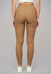 Magas derékú, testhezálló barna leggings sima textúrával, struktúrált varratokkal és szűkített bokával. Fehér sportcipővel párosítva.