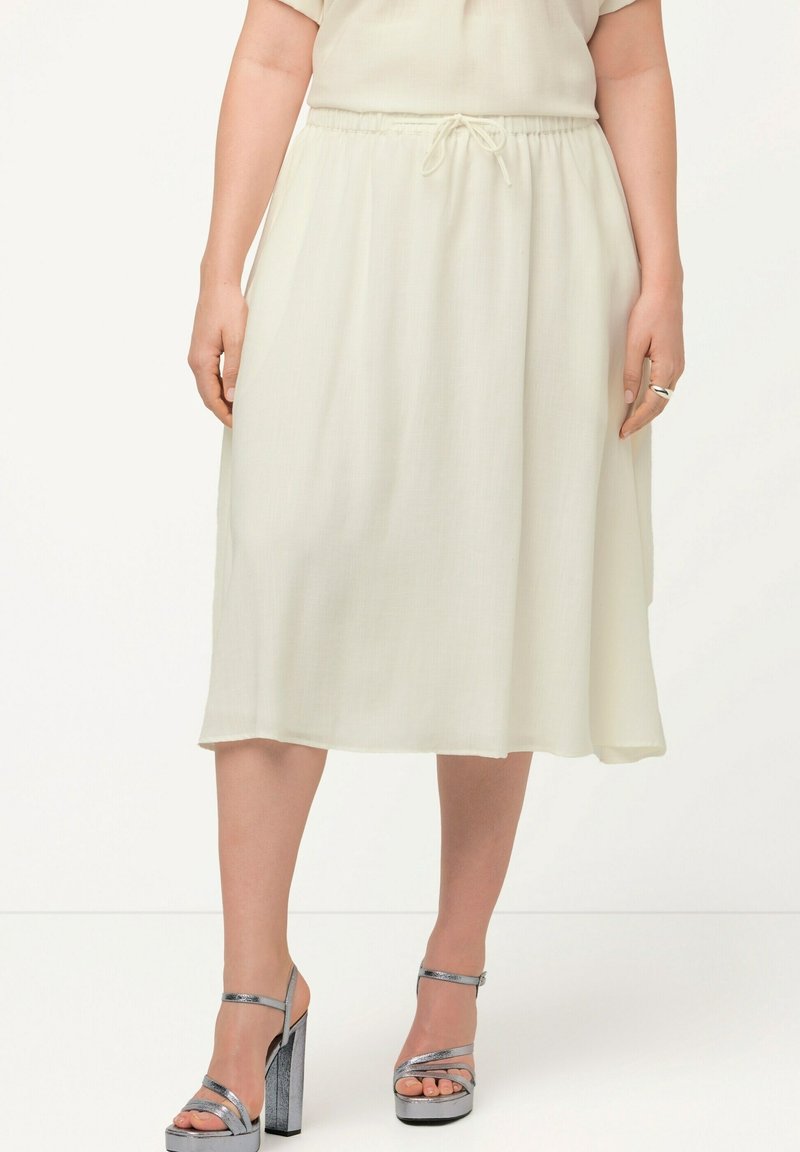 Jupe midi beige clair avec une ceinture élastique et un cordon de serrage ajustable ; texture lisse et design fluide. Associée à des sandales compensées argentées.