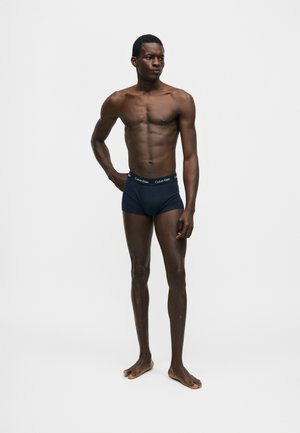 Αθλητικός μελαμψός άνδρας που στέκεται ξυπόλυτος με μπλε σκούρα μποξέρ Calvin Klein, σε απλό ανοιχτό φόντο, με το ένα χέρι στη μέση.