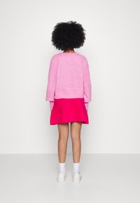 Suéter de punto rosa con mangas largas, combinado con una falda acampanada de un rosa brillante. Se ven calcetines blancos hasta el tobillo y zapatos de color claro.