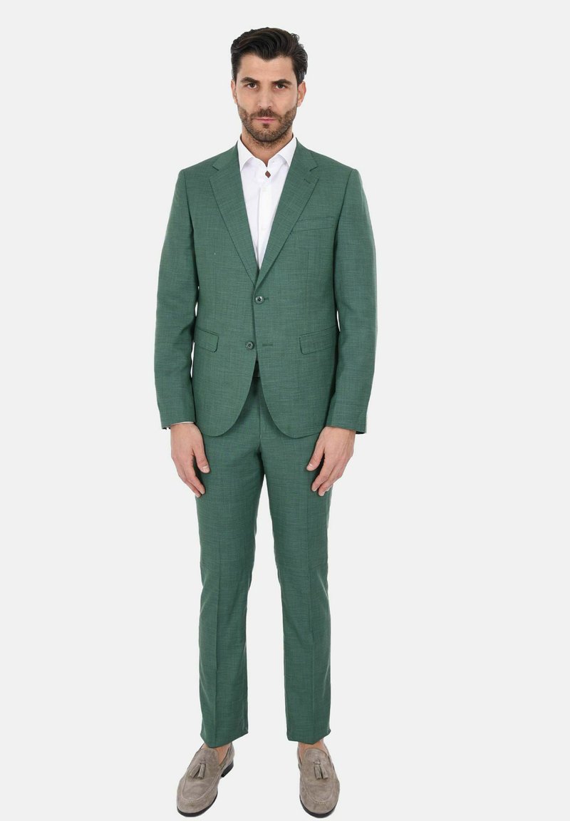 Completo verde con texture, giacca a due bottoni e pantaloni slim-fit, abbinato a una camicia bianca con colletto e mocassini grigi.