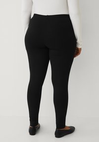 Sorte leggings med en glat tekstur, høj talje og fuld længde, vist fra bagsiden. Båret med flade sorte sko.