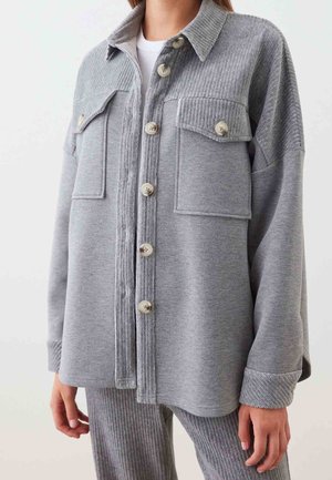 Femme portant une chemise grise à boutons avec des détails côtelés aux épaules et aux poches, associée à un pantalon texturé côtelé assorti.