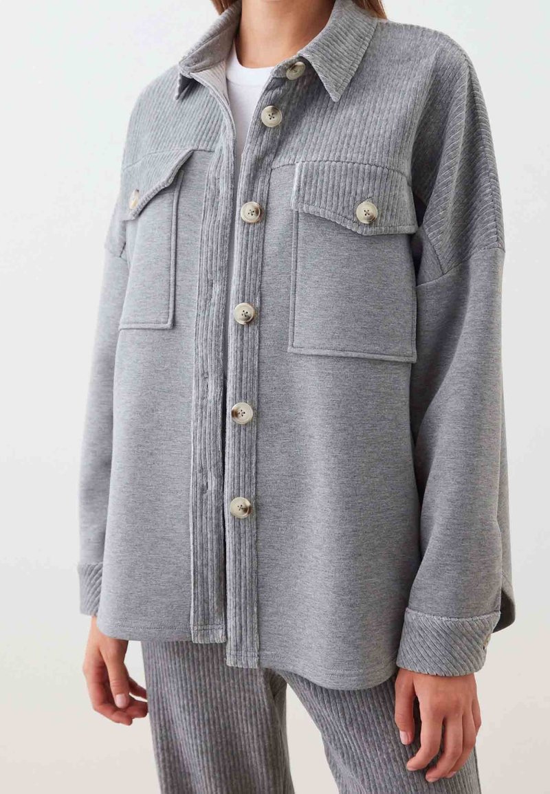 Femme portant une chemise grise à boutons avec des détails côtelés aux épaules et aux poches, associée à un pantalon texturé côtelé assorti.
