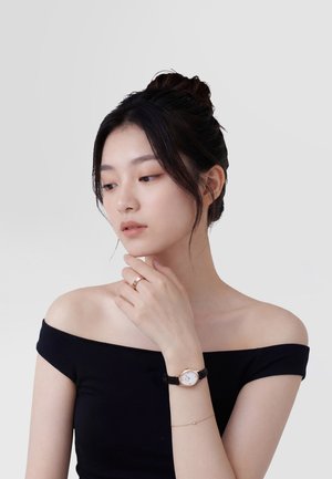 Mlada žena s tamnom kosom u punđi koja nosi crnu bluzu s otvorenim ramenima, sat na zapešću, narukvicu i prsten, zamišljeno gleda u stranu.