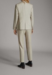 Personne debout, de dos, vêtue d'un costume beige avec un pantalon ajusté et des bottines noires, sur un fond gris uni.