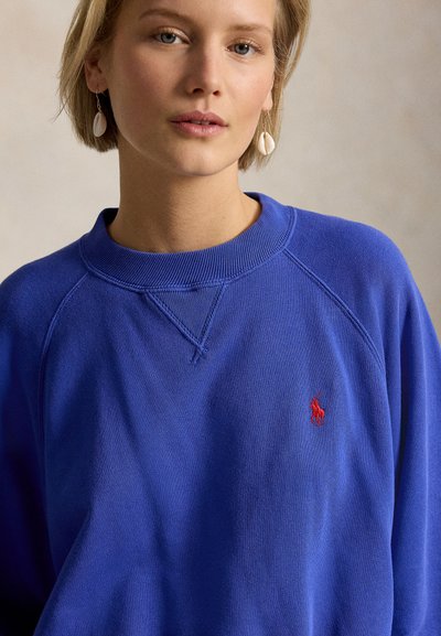 Γυναίκα με κοντά ξανθά μαλλιά, που φορά μπλε φούτερ με κόκκινο κεντημένο λογότυπο και λευκά σκουλαρίκια σε σχήμα κοχυλιού, μπροστά σε μονόχρωμο φόντο.