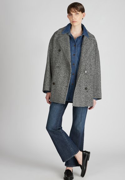 Grijze visgraat dubbelzijdige jas met grote revers, gedragen over een blauwe denim blouse met knopen en donkere flared jeans. Zwarte instapschoenen maken de outfit af.