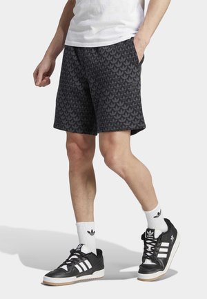 Person iført sorte Adidas-shorts med mønstrede logoer, hvide sokker med logoer og sorte Adidas-sneakers, står med den ene hånd i lommen.