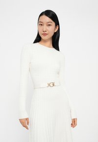 Abito bianco lavorato a maglia con maniche lunghe, caratterizzato da una texture a coste, abbinato a una cintura bianca con una fibbia dorata che mostra la lettera "G."