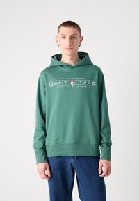 Felpa verde in cotone con tasca a marsupio, cappuccio regolabile con cordino e branding in testo bianco. Indossata con jeans blu a vestibilità ampia.