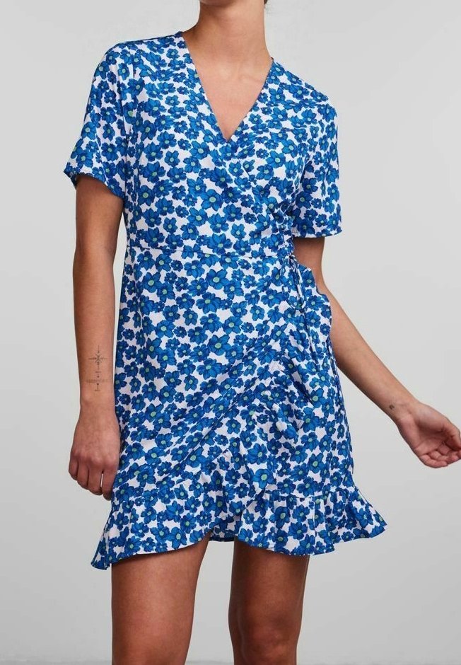 Femme portant une robe portefeuille bleu à fleurs à manches courtes, ourlet à volants et col en V, debout devant un fond gris uni.